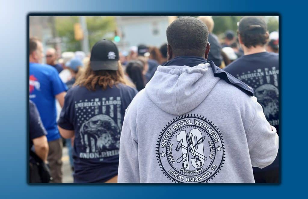 An African American man faces away from the camera, wearing a gray hoodie with the Sheet Metal Workers Local 19 logo on the back, while ahead of him people out of focus march in a parade.